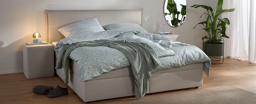 Gemütliches Schlafzimmer mit grauem Boxspringbett, unordentlichen Decken und Kissen, Nachttisch, Lampe, Hausschuhe und Pflanze