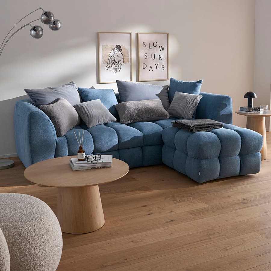 SLOW SUNDAYS – gemütliches Wohnzimmer mit blauer Couch, grauen Kissen, zwei runden Holztischen und Wandbildern