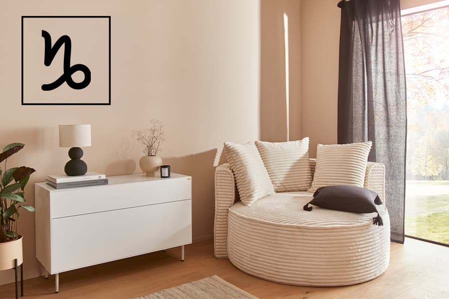 Wohnzimmer mit rundem Sofa und Kissen, weiße Kommode mit Lampe und Pflanzen, Fenster mit schwarzem Vorhang, Wandbild mit Symbol