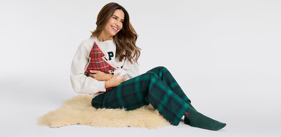 Frau in grün karierten Pyjamahosen und weißem Pullover mit rotem Herz sitzt auf einem Fellteppich.