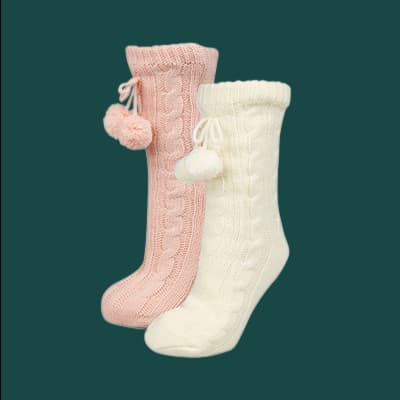 Zwei Paar knielange Socken mit Zopfmuster und Pompons, ein Paar rosa, das andere Paar weiß, vor grünem Hintergrund