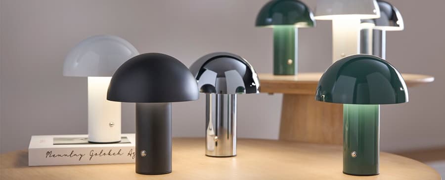 Sechs Tischlampen in verschiedenen Farben und Oberflächen auf einem Holztisch, einige auf einem Buch mit handgeschriebenem Text