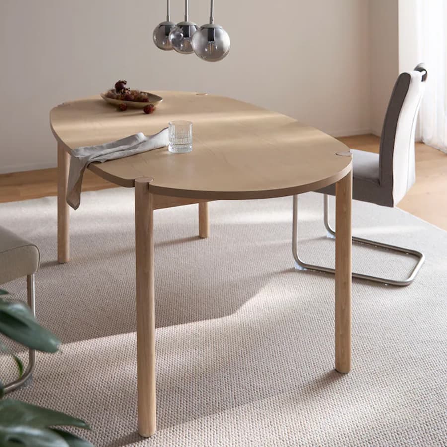 Ovaler Holztisch mit Glas und Schale auf heller Teppichfläche, daneben grauer Stuhl unter drei hängenden Lampen