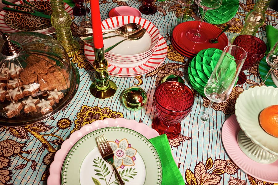 Bunt gedeckter Tisch mit Tellern, Gläsern, Besteck, Kerzen und Gebäck in Glasglocke, grüne Servietten, Mandarine auf Teller