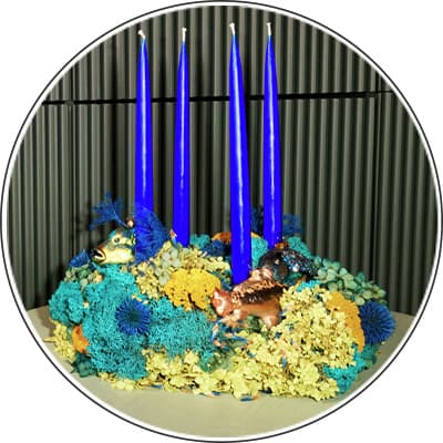 Vier blaue Kerzen in einem Kranz aus bunten Blumen mit Fisch- und Tierfiguren auf einem Tisch vor einem gestreiften Hintergrund