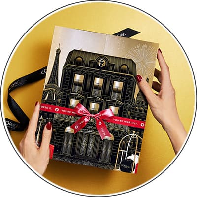 Person hält L'Oréal Adventskalender mit roter Schleife und dem Text "You're worth it" vor einem goldenen Hintergrund