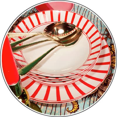Gestapelte Teller mit roten Streifen, darauf goldene Löffel und Gabeln, neben einem roten Kerzenhalter auf gemustertem Untergrund