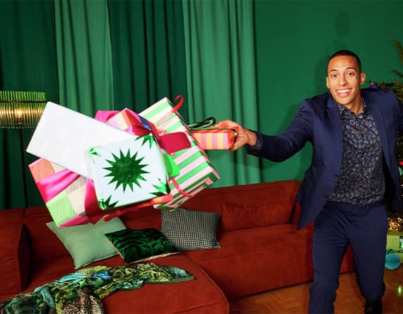 Mann im Anzug hält viele bunte Geschenkboxen vor einem roten Sofa und grünen Vorhängen