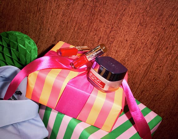 Geschenk mit pink-gelbem Streifenpapier und pinker Schleife, darauf Lippenstift, Parfümflakon und Creme.