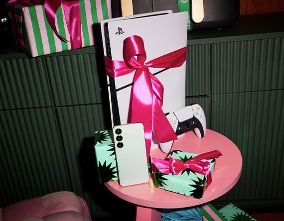 PlayStation 5 mit roter Schleife, Controller, Smartphone und Geschenke auf rosa Tisch vor grünem Hintergrund