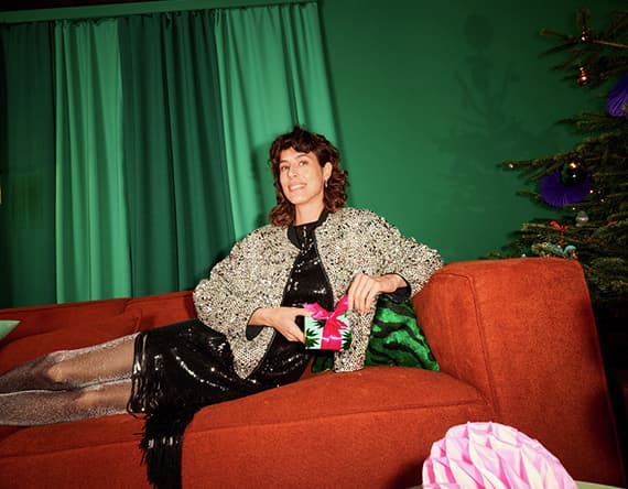 Frau in schwarzem Kleid und silbernem Mantel sitzt auf rotem Sofa, hält Geschenkbox. Hintergrund mit grünem Vorhang und Weihnachtsbaum