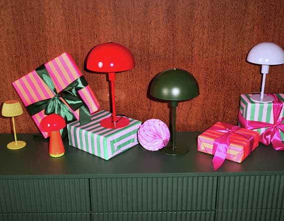 Bunte Tischlampen und gestreifte Geschenkboxen mit Schleifen auf grünem Möbelstück vor Holzpaneel-Hintergrund