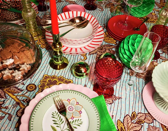 Bunt gedeckter Tisch mit gemustertem Geschirr, Gläsern, Kerzen, Besteck und einer Kuchenglocke auf blumiger Tischdecke