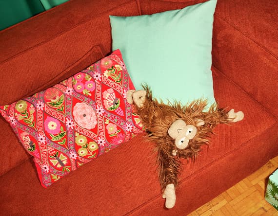 Stoffaffe mit freundlichem Gesicht liegt auf rotem Sofa zwischen buntem und türkisfarbenem Kissen