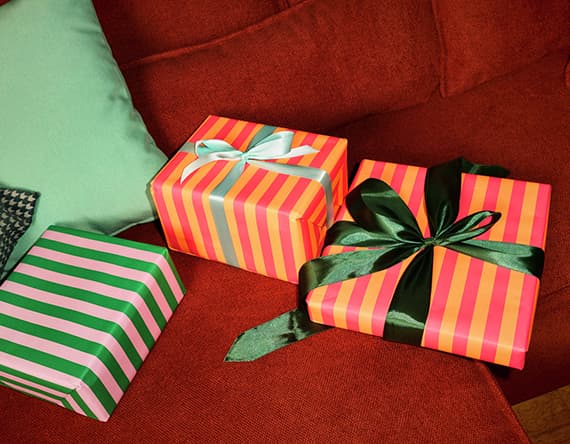 Drei farbenfrohe, gestreifte Geschenke mit Schleifen auf rotem Sofa, grünes Kissen im Hintergrund