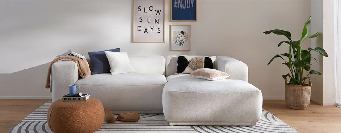 Wohnzimmer mit weißem Sofa und Hocker, dekoriert mit Kissen und Decke. Wandbilder mit "SLOW SUNDAYS" und "ENJOY". Pflanze im Korb.