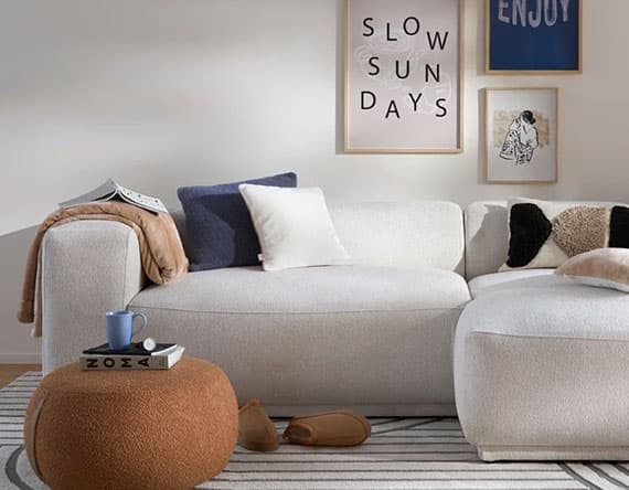 SLOW SUN DAYS – Gemütliches Wohnzimmer mit weißem Sofa, Kissen, Decken, Hocker, Tasse, Buch, und Wandbildern.