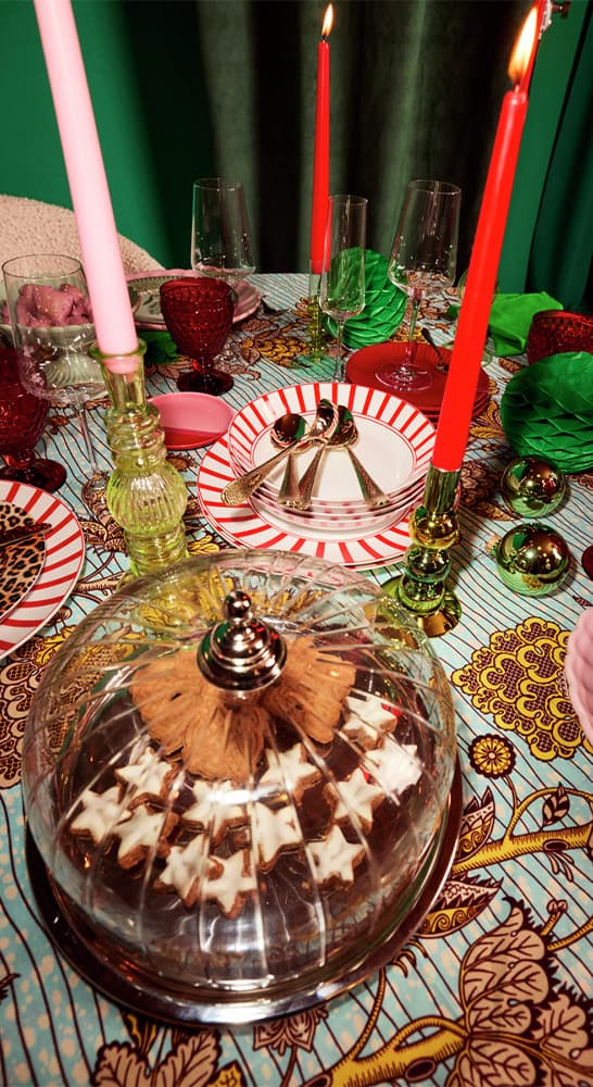 Tischgedeck mit Kerzen, Rotweingläsern, Besteck und Teller. Gebäck in Sternform unter Glasglocke auf gemustertem Tischtuch.