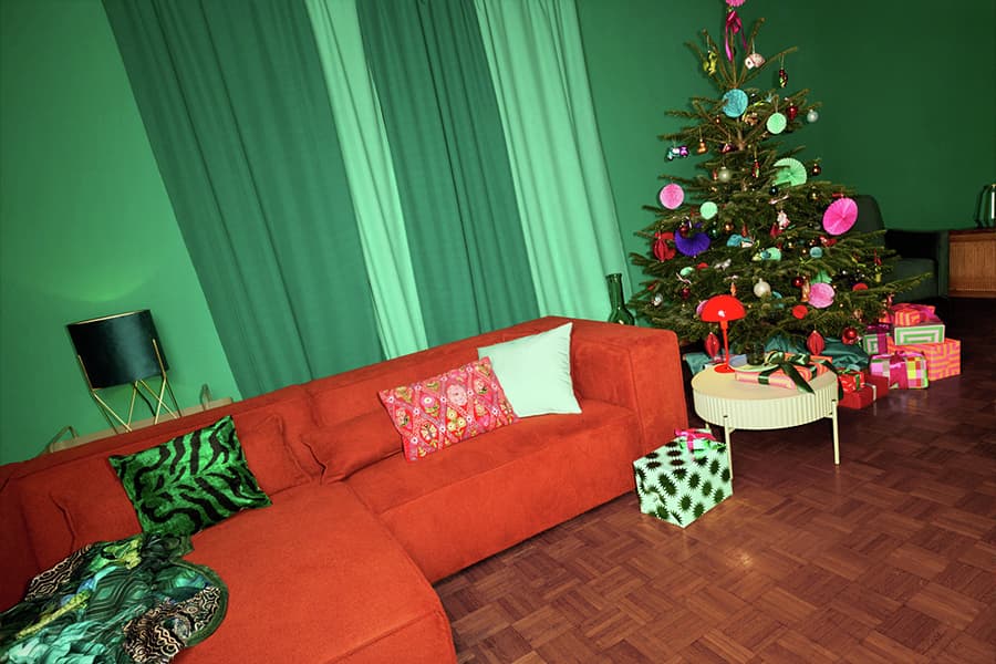 Wohnzimmer mit orangefarbenem Sofa, grüne Kissen, Weihnachtsbaum mit bunten Kugeln, Geschenke auf dem Boden und Tisch