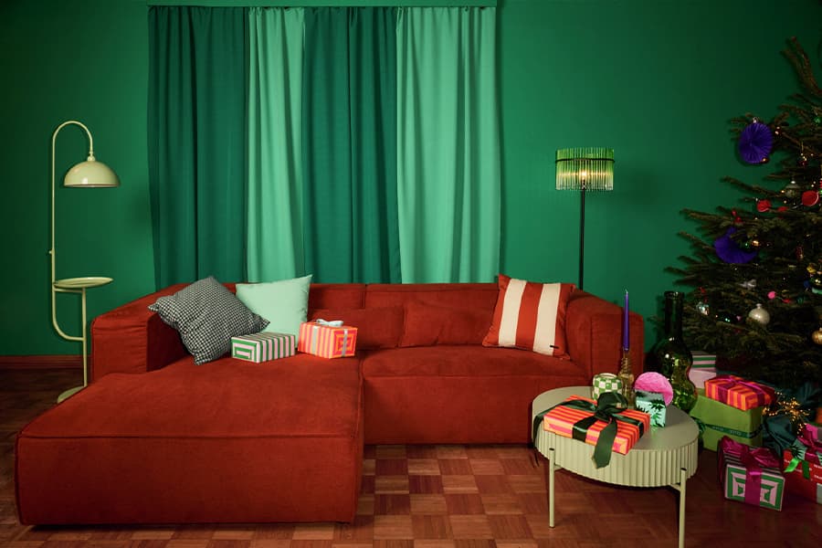 Wohnzimmer mit orangefarbenem Sofa, grüne Kissen, Weihnachtsbaum mit bunten Kugeln, Geschenke auf dem Boden und Tisch
