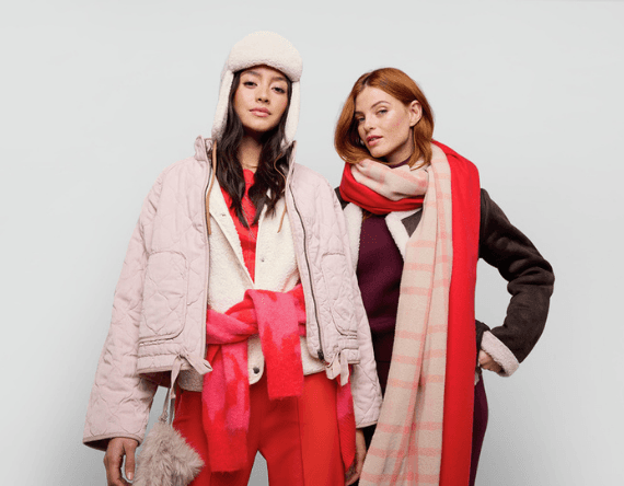 Zwei Frauen in Winterkleidung mit warmen Mänteln, Schals und Mützen stehen nebeneinander vor grauem Hintergrund