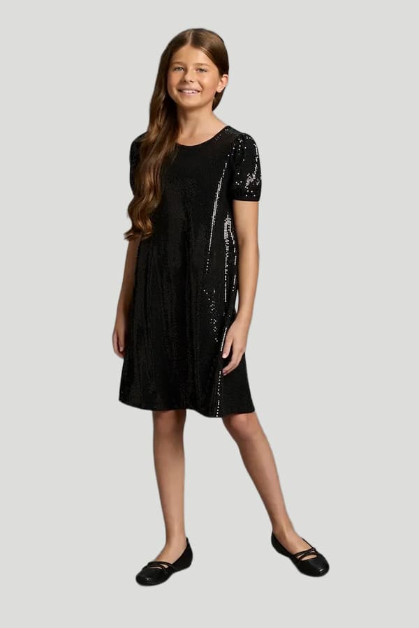 Mädchen in schwarzem Paillettenkleid mit kurzen Ärmeln und schwarzen Ballerinas steht vor neutralem Hintergrund.
