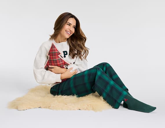 Frau sitzt auf einem Teppich und hält ein rotes Kissen mit Tannenbaum-Motiv, trägt weiße Jacke und grün karierten Hose.