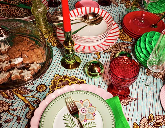 Bunter festlicher Tisch mit Teller, Besteck, roten und grünen Gläsern, Kerzen und einem Glasdeckel voller Kekse.