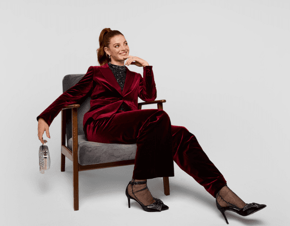 Frau sitzt in grauem Stuhl, trägt roten Anzug und schwarze High Heels, hält silberne Handtasche
