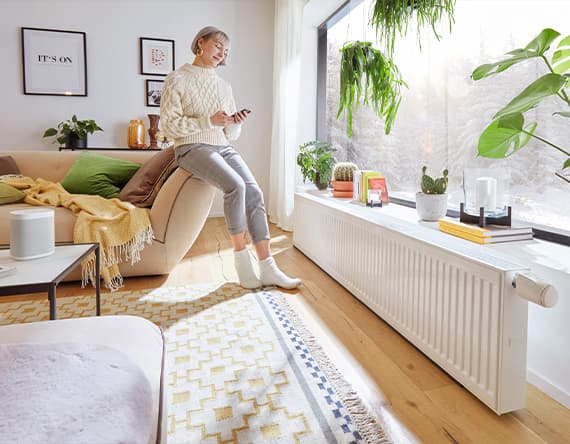 Person in gemütlichem Wohnzimmer, sitzt auf Sofa, hält ein Handy. Große Fenster, Pflanzen und Bücher auf Fensterbank.