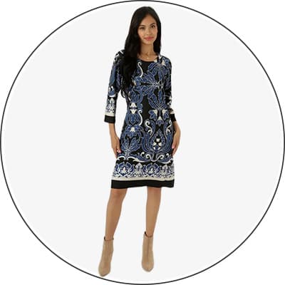 Frau trägt knielanges Kleid mit blauem Blumenmuster und langen Ärmeln, steht mit einer Hand an der Hüfte und trägt beigefarbene Stiefeletten.