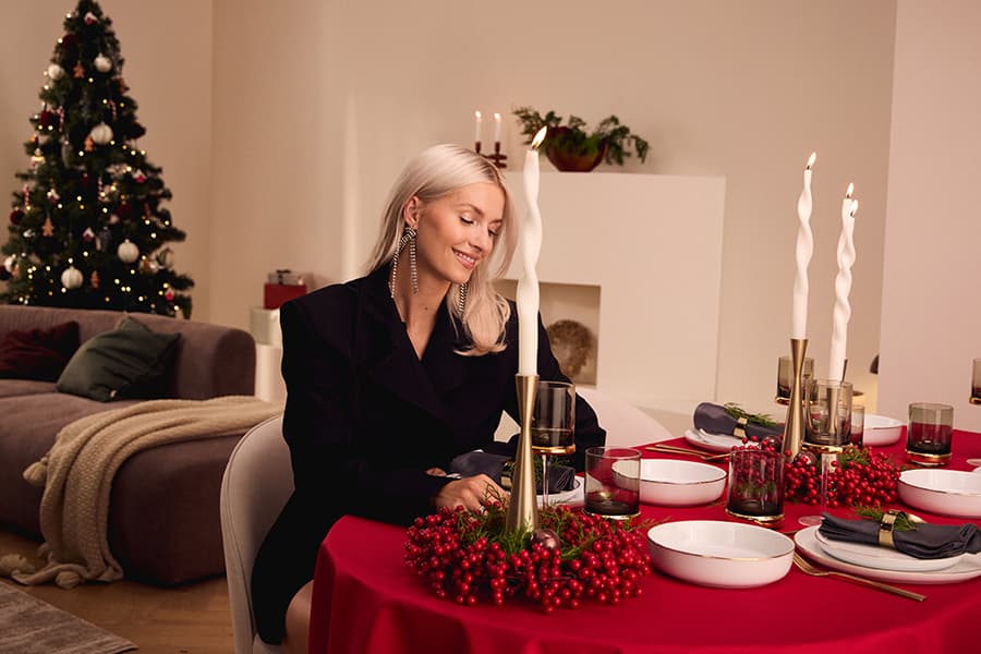 Frau mit langen, blonden Haaren sitzt an einem festlich gedeckten Tisch mit roten Beeren, Kerzen und Geschirr, im Hintergrund ein geschmückter Weihnachtsbaum