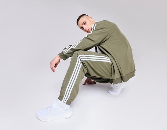 Mann in olivgrünem Trainingsanzug mit weißen Streifen und weißen Sneakers hockt vor grauem Hintergrund.