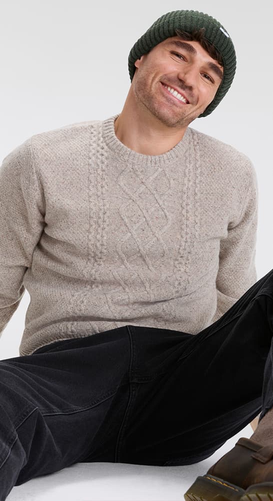 Mann trägt beigen Strickpullover und dunkelgraue Hose, sitzt auf dem Boden mit grünem Strickmütze.