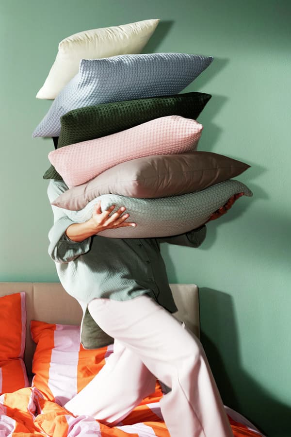 Person trägt sechs bunte Kissen, steht neben einem Bett mit orange-gestreifter Bettwäsche vor grünem Hintergrund.