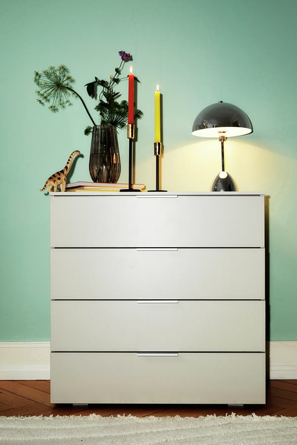 Weiße Kommode vor mintgrüner Wand, dekoriert mit Vase, Kerzen und Lampe. Teppich auf Holzboden.