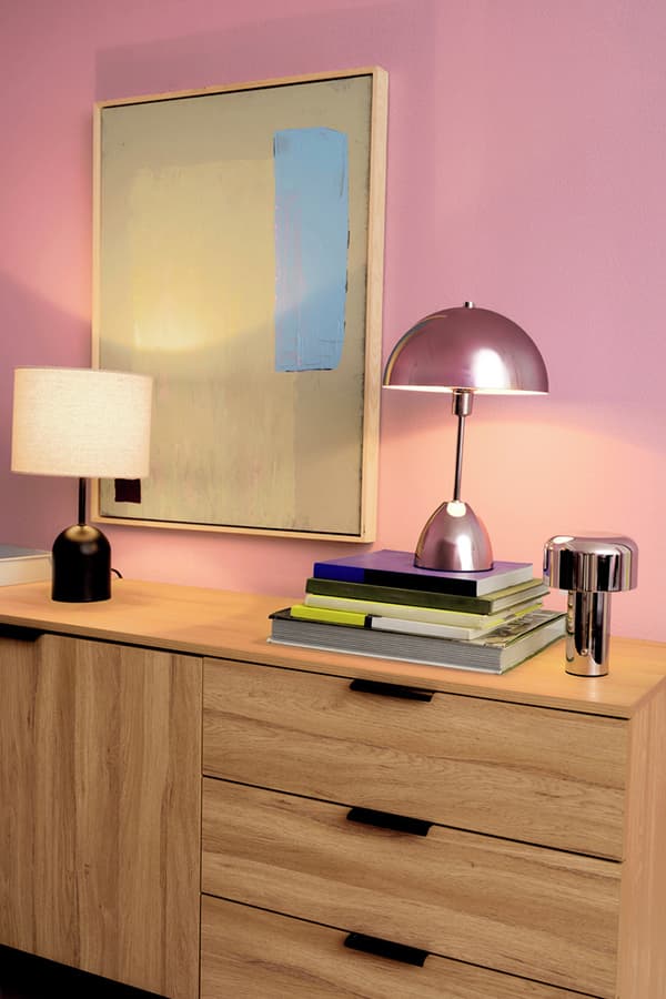 Holzkommode mit zwei Lampen, Büchern und einem abstrakten Bild an der rosa Wand