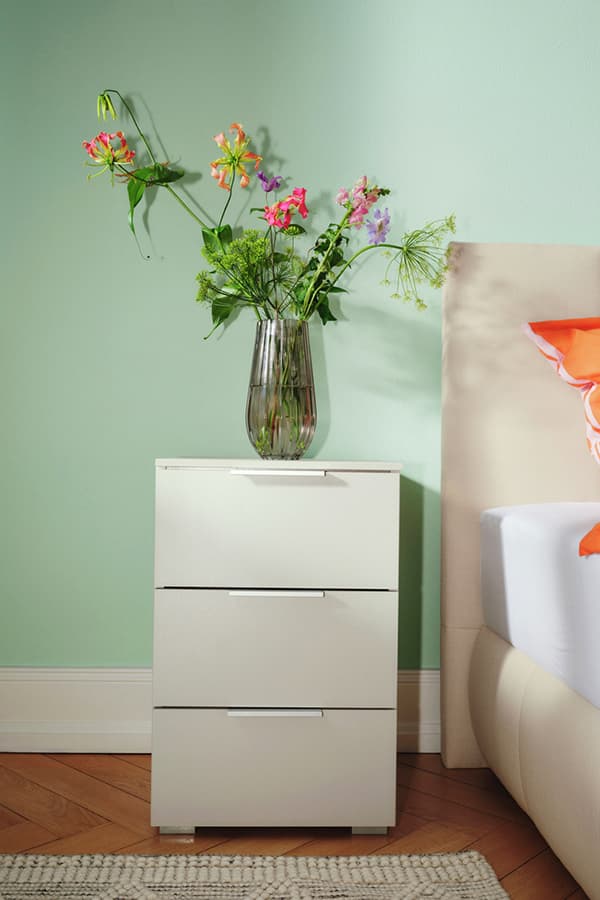 Weiße Kommode mit drei Schubladen, darauf eine Vase mit bunten Blumen, neben einem Bett mit orangefarbenem Kissen.