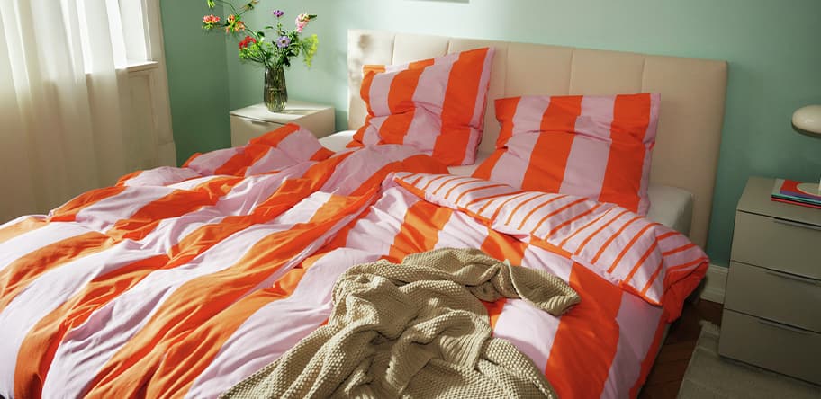 Bett mit orange-weiß gestreifter Bettwäsche, beiger Decke und Vase mit Blumen auf Nachttisch.