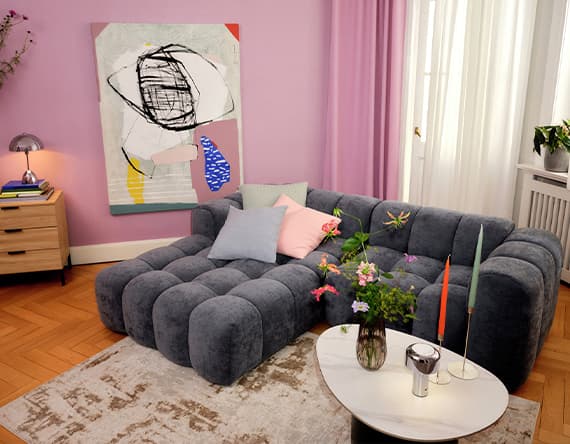 Wohnzimmer mit grauem Sofa, abstraktem Kunstwerk an der Wand, Tisch mit Blumen und Kerzen, lila Wandfarbe und Vorhänge.