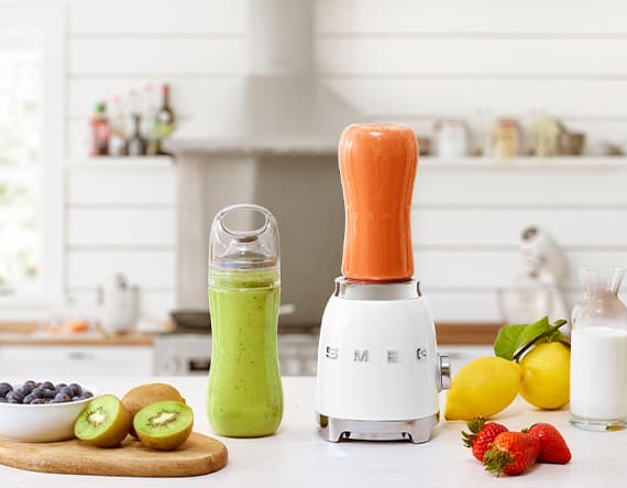Ein weißer Smeg-Standmixer mit orangefarbenem Smoothie, daneben grüner Smoothie, Früchte und Milch in einer Küche.