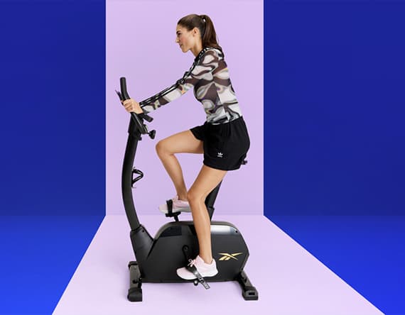 Frau auf einem Heimtrainer in sportlicher Kleidung, vor einem farbigen Hintergrund.