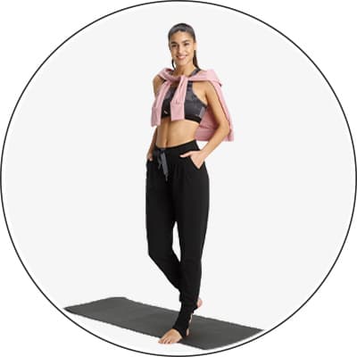 Frau in Sport-BH und Jogginghose steht auf einer Yogamatte, mit Pullover über den Schultern.