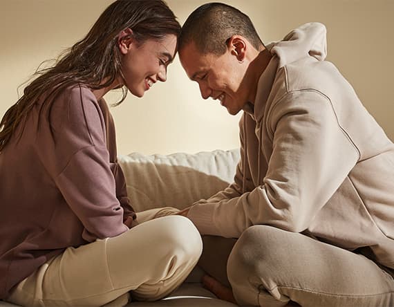 Zwei Personen sitzen gegenüber und lehnen ihre Stirn aneinander. Beide tragen gemütliche Kleidung.