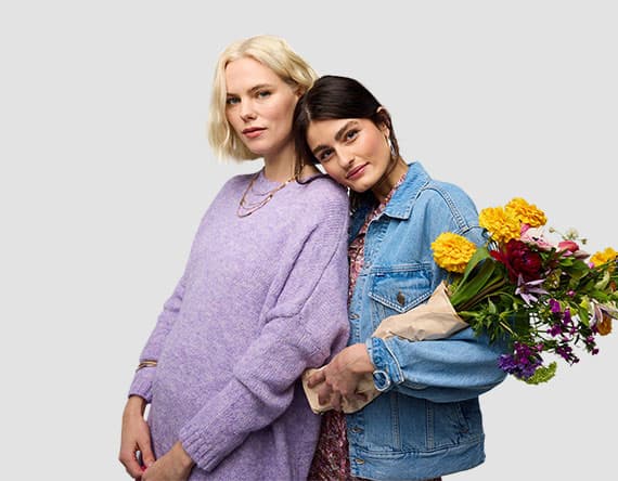 Zwei Personen stehen nebeneinander, eine trägt einen lila Pullover, die andere eine Jeansjacke und hält einen Blumenstrauß.