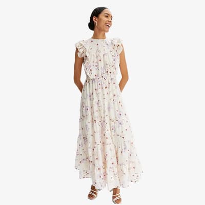 Frau in langem, weißem Kleid mit buntem Blumenmuster und Rüschenärmeln, steht mit Händen in den Taschen.