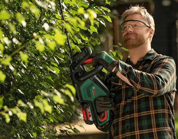 Mann in kariertem Hemd schneidet mit grünem Elektro-Heckenschneider einen Busch.