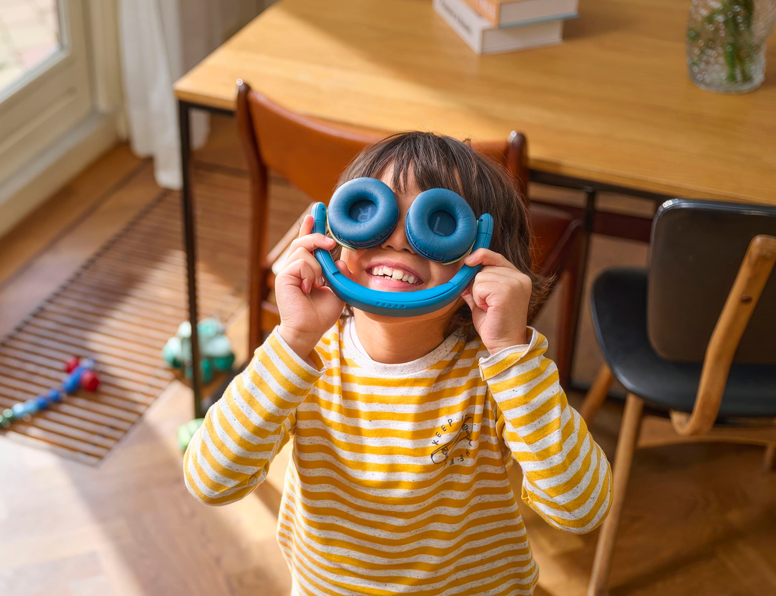 Kind in gelb-weiß gestreiftem Pullover hält ein Spielzeug vor das Gesicht in einem hellen Raum mit Holzmöbeln.