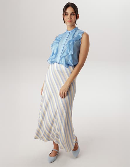 Frau trägt eine blaue Bluse mit Rüschen und einen gestreiften langen Rock. Sie steht leicht zur Seite gedreht.