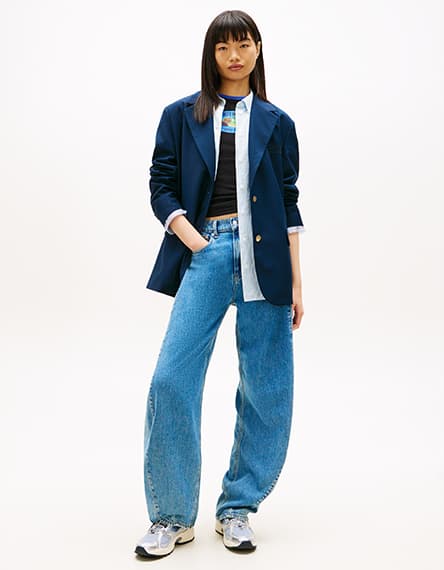 Frau in dunkelblauem Blazer und weiten Jeans mit den Händen in den Taschen. Sie trägt sportliche Turnschuhe.